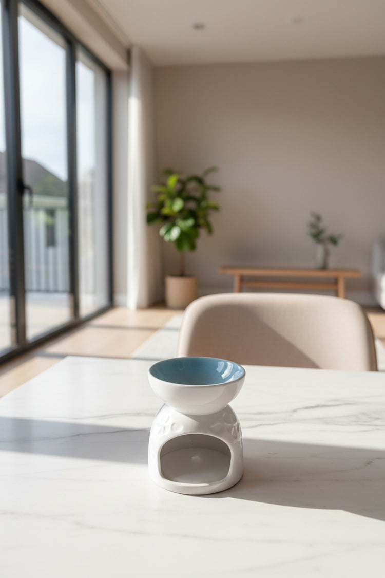A white ceramic oil burner with a light blue glazed interior, sitting on a gray wooden table in a living room setting.