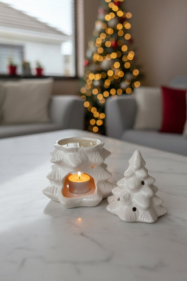 A white ceramic Christmas tree-shaped candle holder with a tiered, textured design, shown with its lid placed beside it on a gray wooden table. The holder is lit from within, casting a warm orange glow.