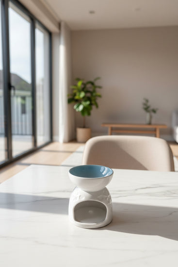 A white ceramic oil burner with a light blue glazed interior, sitting on a gray wooden table in a living room setting.